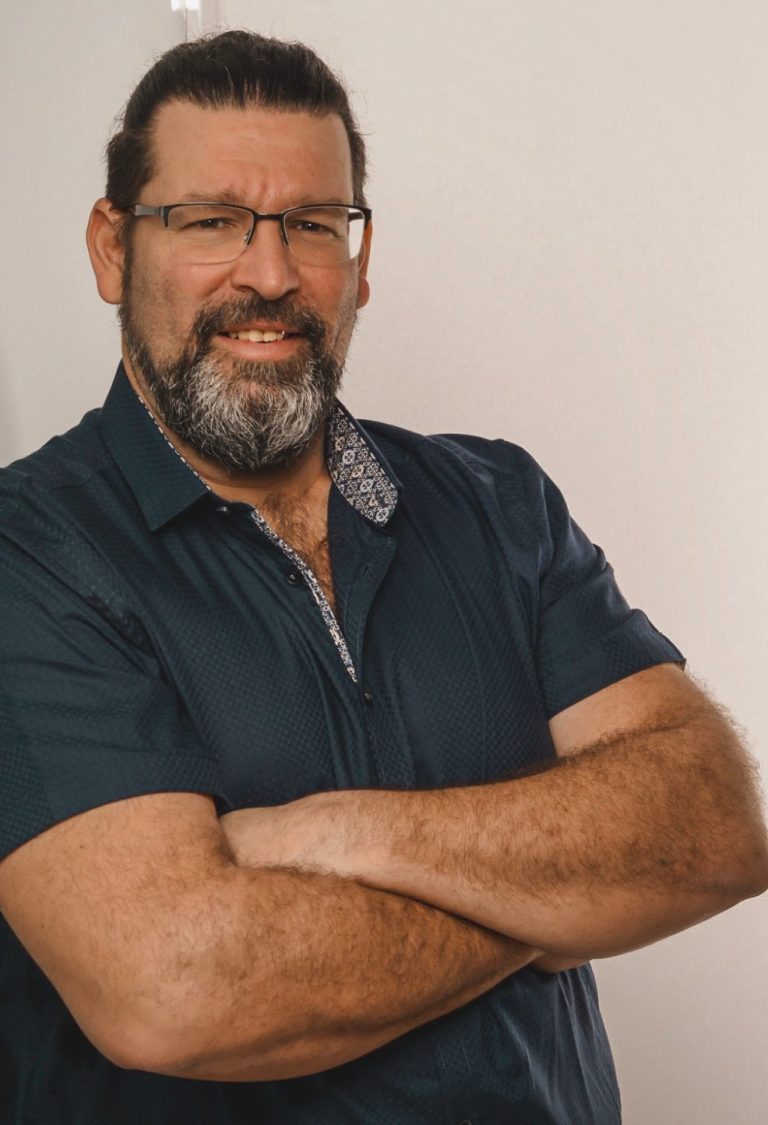 Mann mit Brille und Bart, lächelt mit verschränkten Armen vor einer hellen Wand.